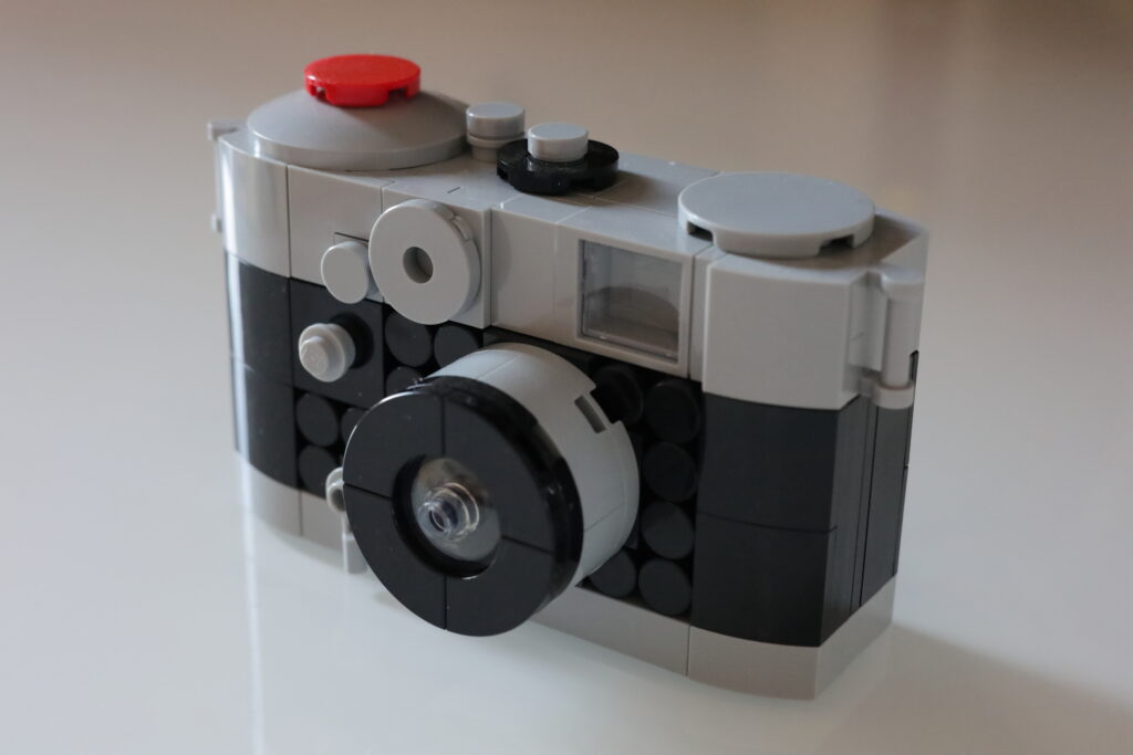 Das Bild zeigt eine Kamera aus Lego. Sie ist oben und unten grau und in der Mitte schwarz. der Auslöser ist rot.